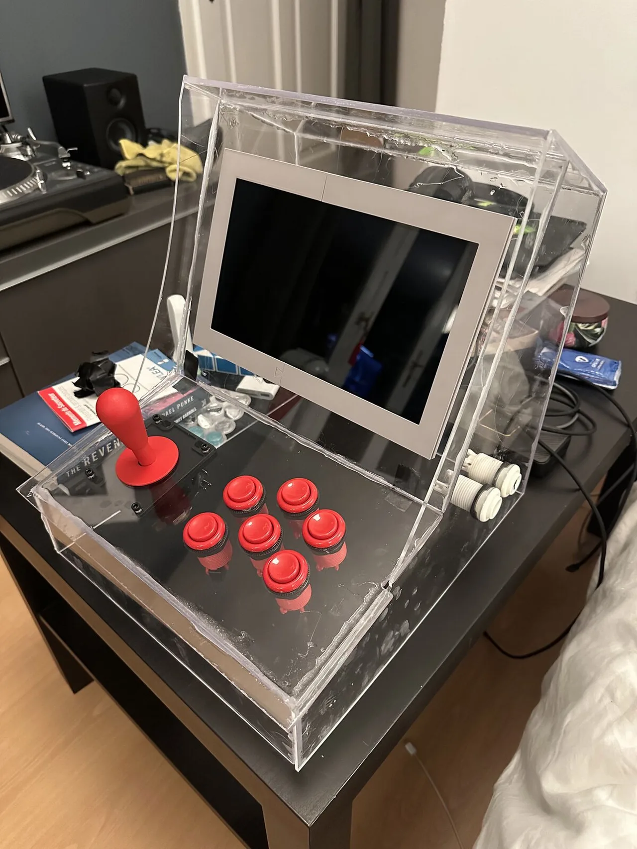A small clear plastic arcade cabinet with a screen, a red joystick, six red buttons and two white buttons on the side