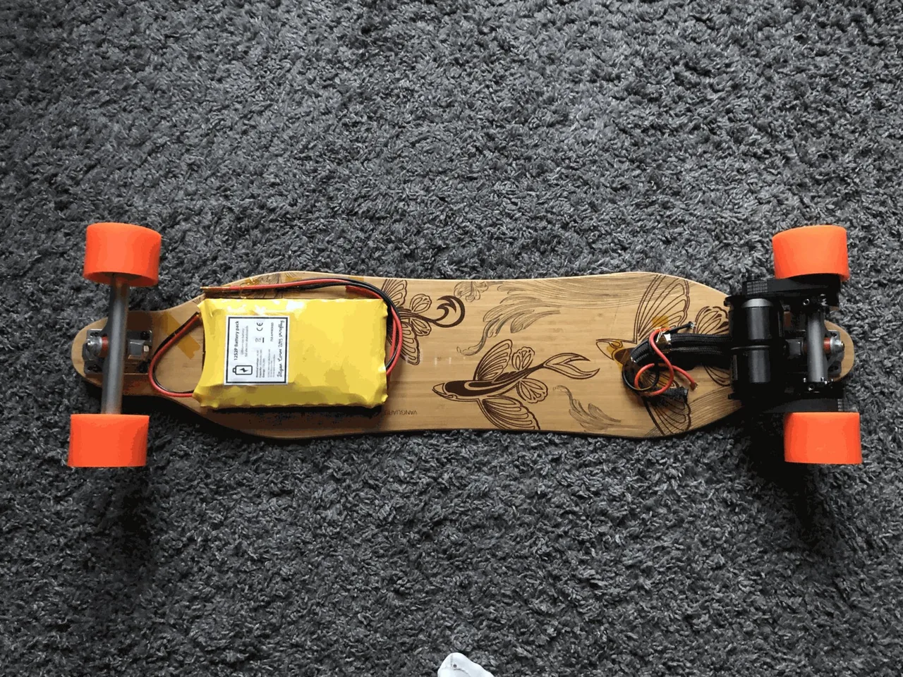 A bamboo longboard seen from below, with a yellow 12S2P battery pack on the deck and a motor on the rear truck