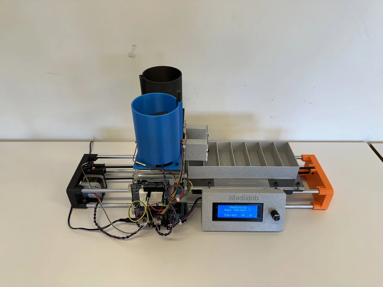 The Medistrib prototype: two pill hoppers, a belt-driven tray with seven compartments, an Arduino stack and an LCD reading Quel patient