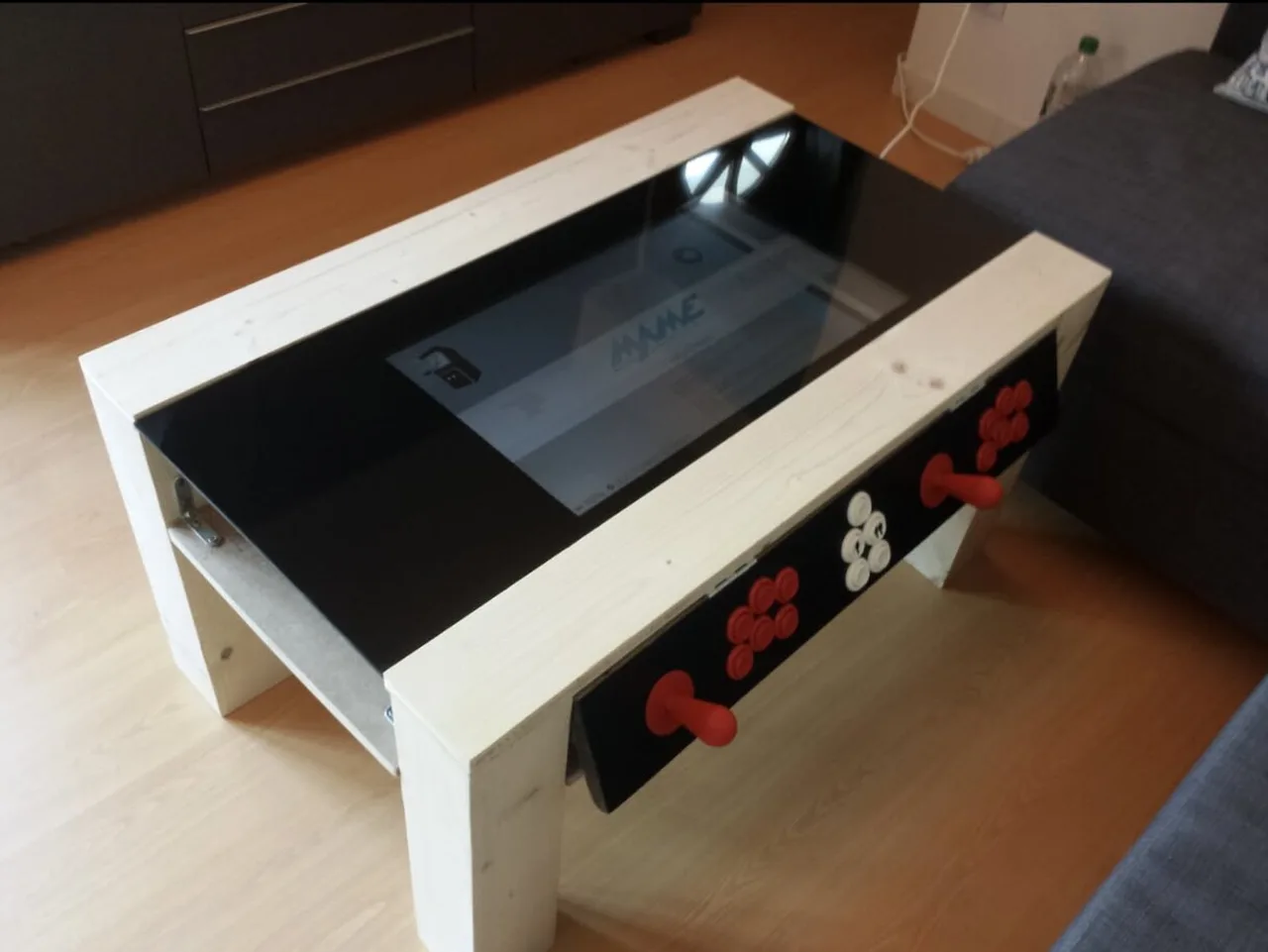 A white wooden coffee table with a screen under black glass and a two-player control panel with red joysticks and buttons