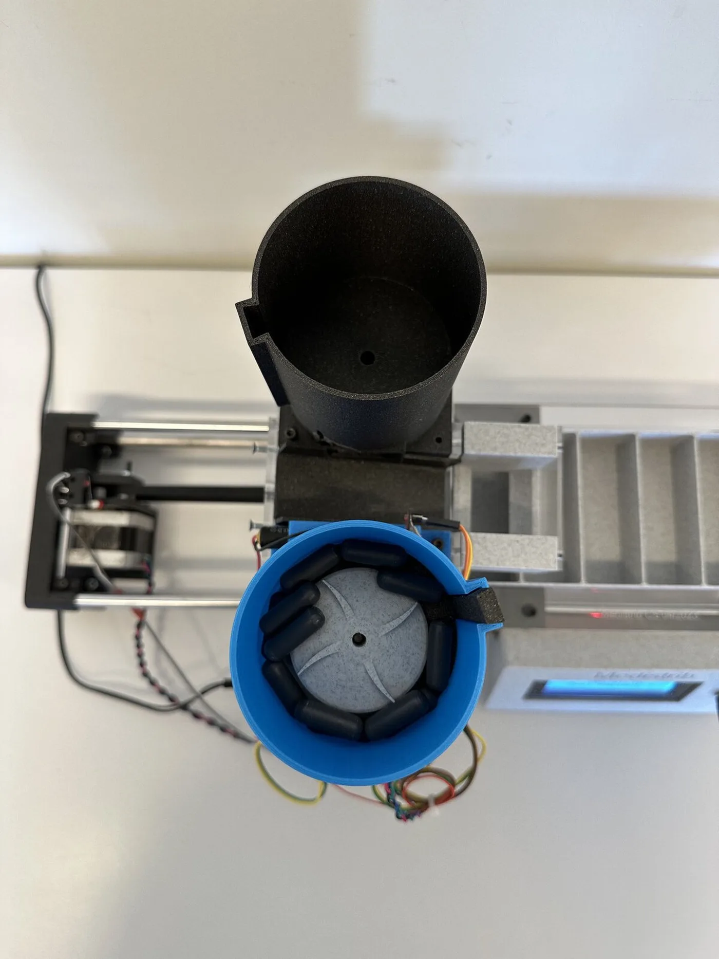 Top view: two hoppers, the blue one holding dark capsules around a slotted disc, above the compartment tray